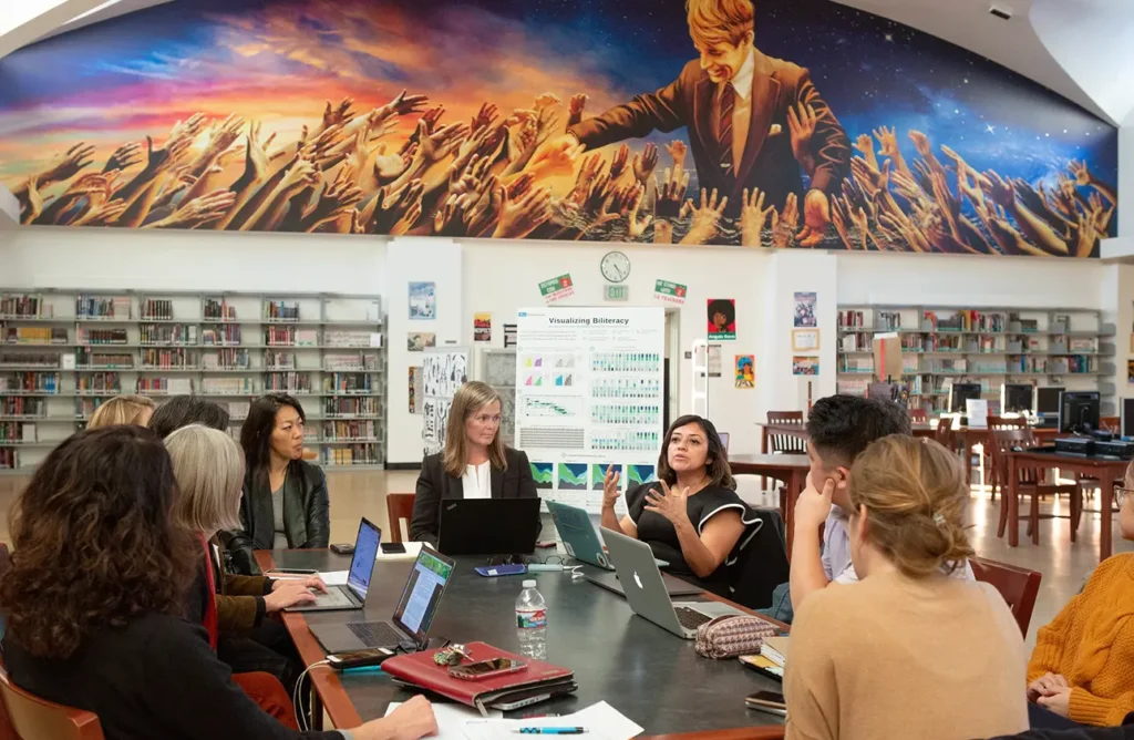 Educators gathered around a library table discussing literacy data and instructional strategies, with a large mural of Robert F. Kennedy and outstretched hands in the background.
