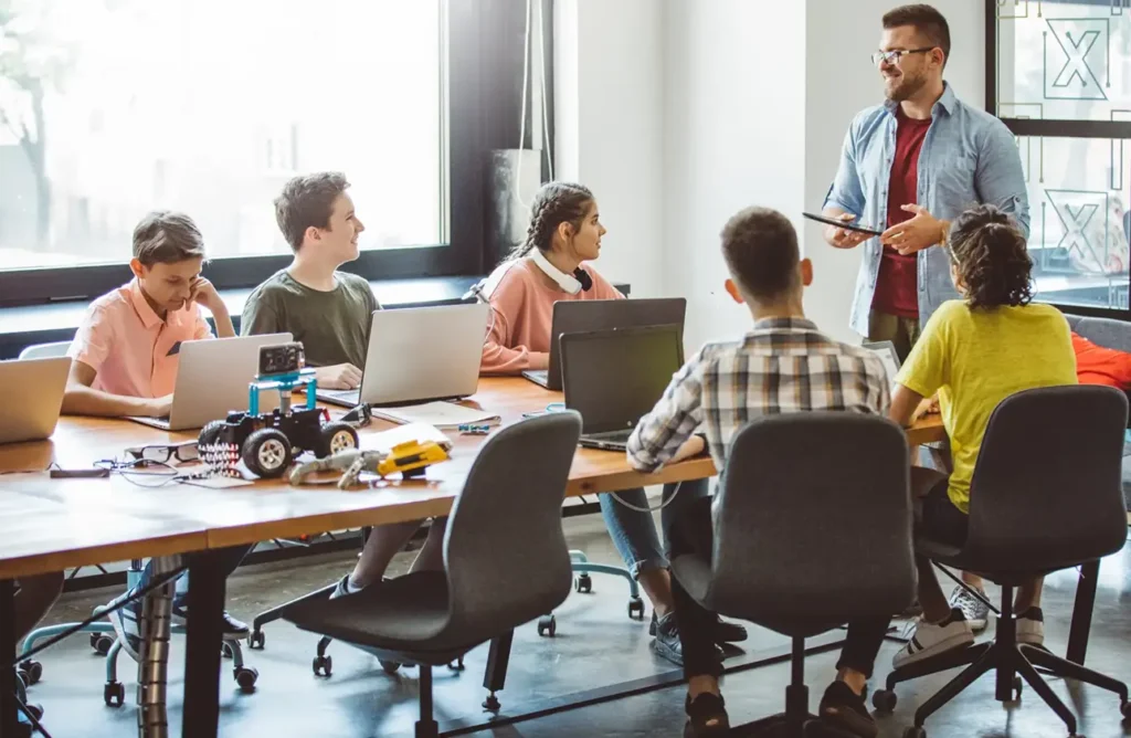 Teacher leading a classroom discussion with students working on laptops and robotics projects around a large table.