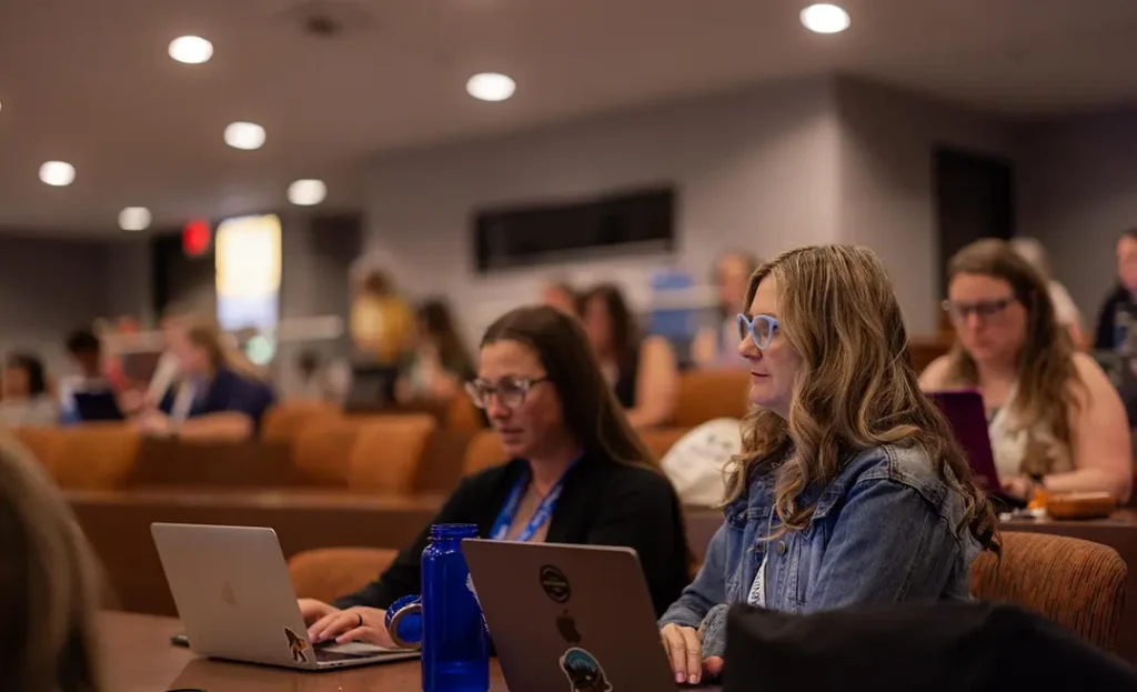 Educators sitting in a lecture hall taking notes on laptops during a professional learning session.