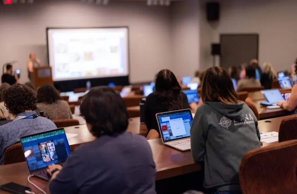 Conference attendees sit with laptops open, following along with a speaker’s presentation projected on a large screen at the front of the room.