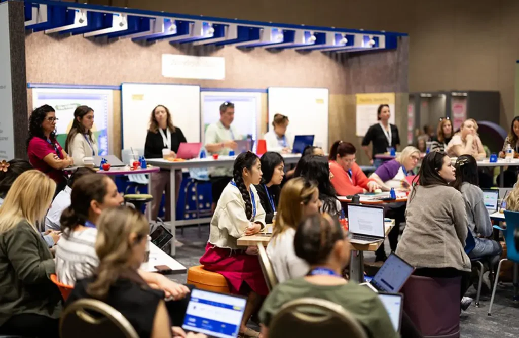 Educators gathered for a collaborative session at a conference, actively listening and taking notes on laptops in a modern learning space.