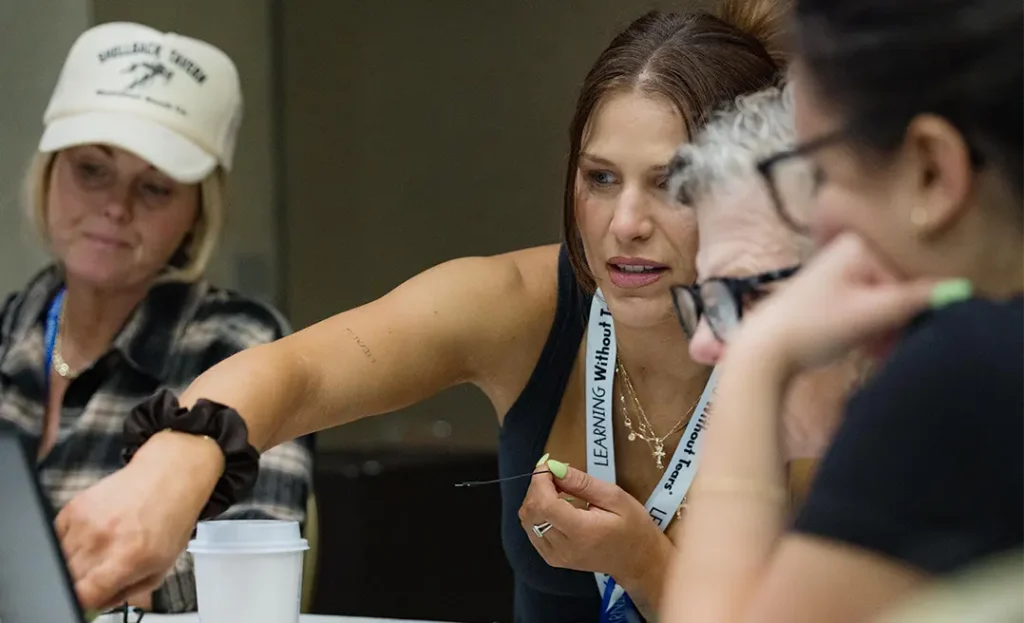 Educators collaborate during a hands-on workshop, with one participant pointing to a laptop screen while discussing ideas with her group.